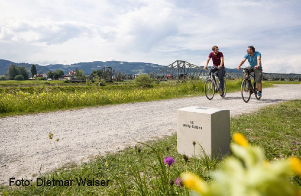 Zwei Männer fahren auf einem Kiesweg Fahrrad, im Vordergrund steht ein Grenzstein.
