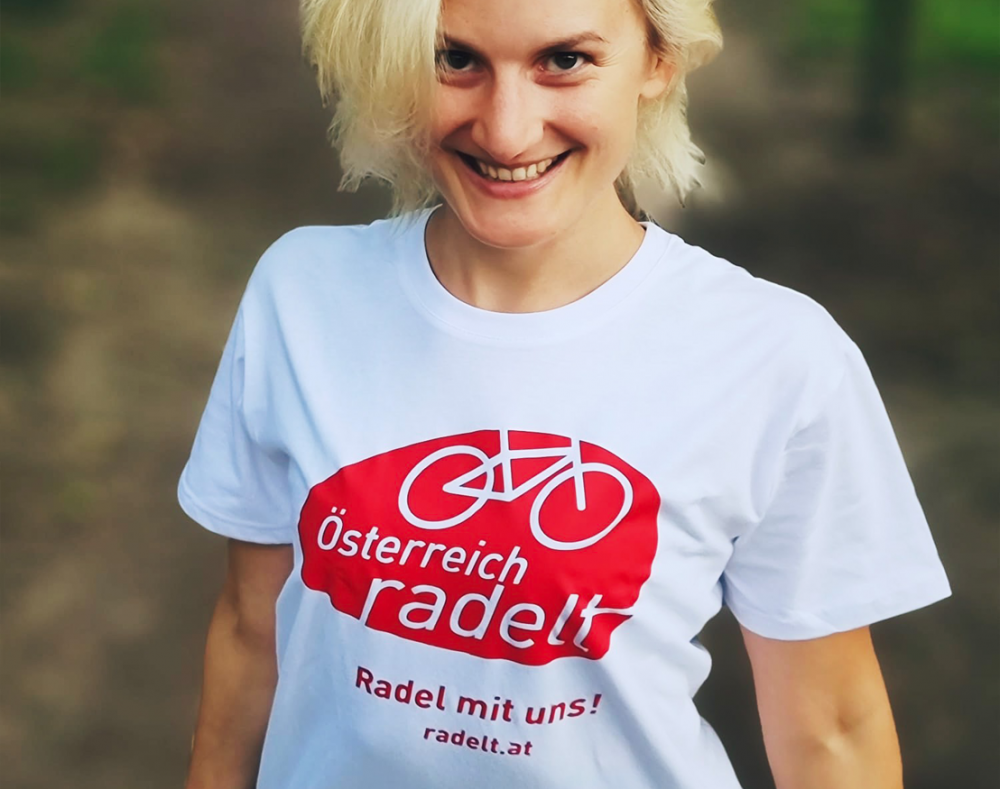 Lächelnde Frau mit weißem T-Shirt auf dem das Österreich radelt Logo gedruckt ist.