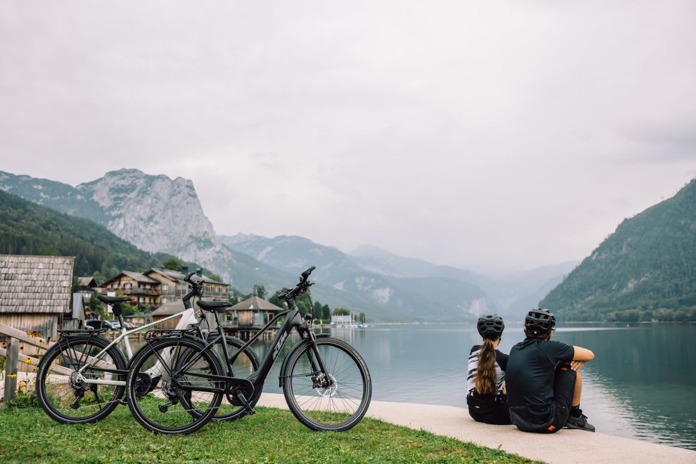 2 Räder stehen neben See und Bergen, Radler und Ralderin sitzen und schauen in die Ferne